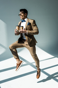 stylish mixed race man in suit holding coffee to go and jumping on grey with sunlight