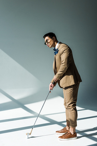 stylish mixed race man in suit holding golf club on grey with sunlight