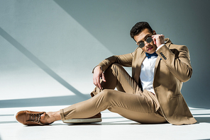 stylish mixed race man in suit and sunglasses sitting and posing on grey with sunlight and copy space