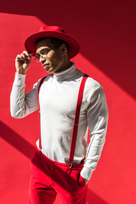fashionable mixed race man in hat and suspenders holding glasses while posing on red