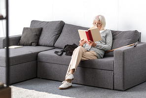 senior woman in glasses sitting on sofa and reading book in living room