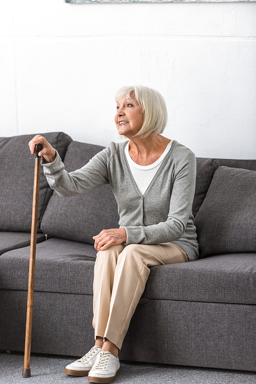 smiling senior woman with cane sitting on sofa and looking away in living room