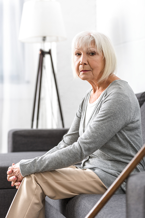 pensive senior woman with wooden cane sitting on sofa in living room