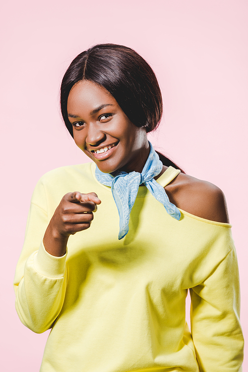 smiling african american woman in yellow pullover and scarf pointing with finger isolated on pink