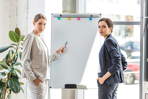 two businesswomen in formal wear standing near flipchart in office
