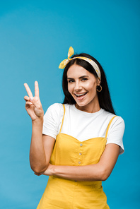 happy young woman in headband showing peace sign  isolated on blue