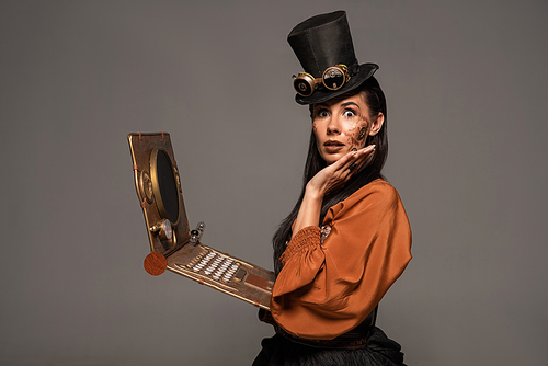 shocked woman in top hat with goggles using steampunk laptop isolated on grey