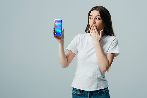 shocked beautiful girl in white t-shirt showing smartphone with booking app isolated on grey