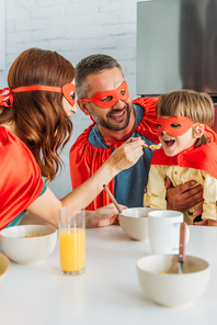 cheerful family in costumes of superheroes having breakfast, while mother feeding son with flakes