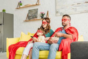 family in costumes of superheroes sitting on sofa, eating popcorn and watching tv