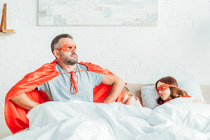 serious father in costume of superhero sitting in bed near sleeping family, holding hands on hips and looking away