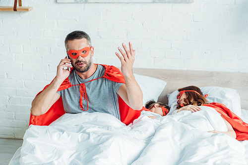 man in superhero costume talking on smartphone and gesturing while sitting in bed near sleeping family