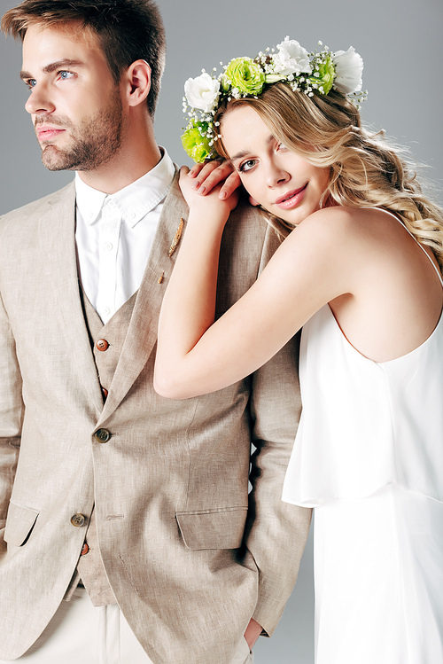 bride in wedding dress and wreath hugging handsome bridegroom in suit isolated on grey