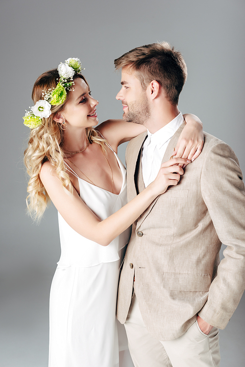 handsome bridegroom in suit hugging with bride in wedding dress and wreath isolated on grey