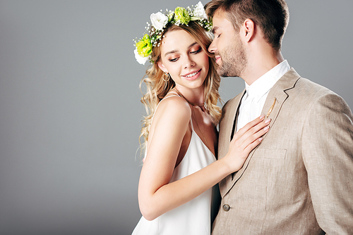 handsome bridegroom in suit hugging with bride in wedding dress and wreath isolated on grey