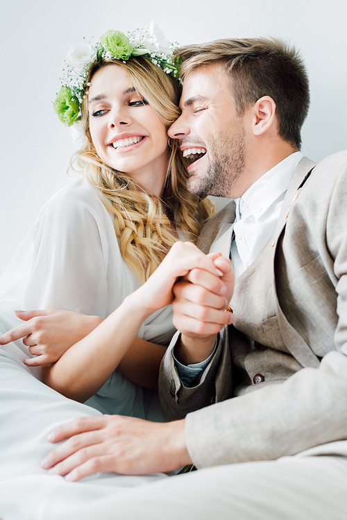 attractive bride and handsome bridegroom holding hands and looking away isolated on grey