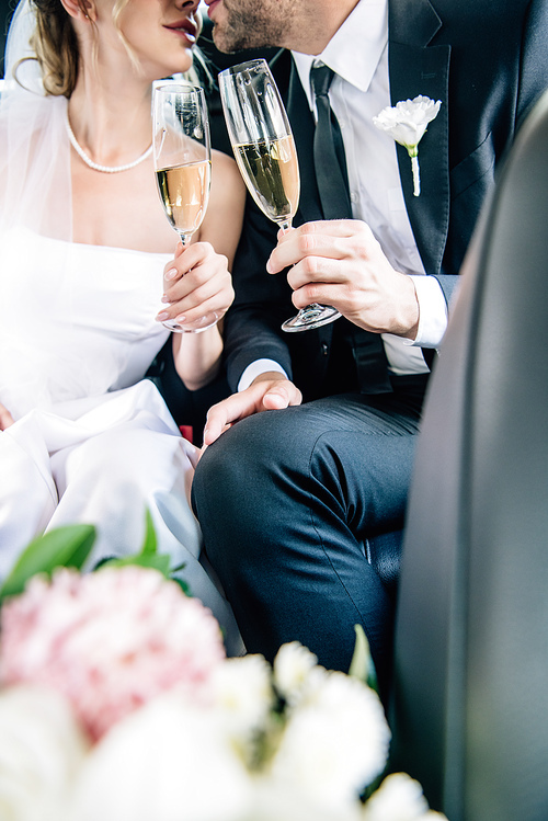 cropped view of bride and bridegroom kissing and clinking with champagne glasses