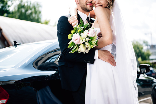 cropped view of bridegroom in suit hugging bride in wedding dress with bouquet