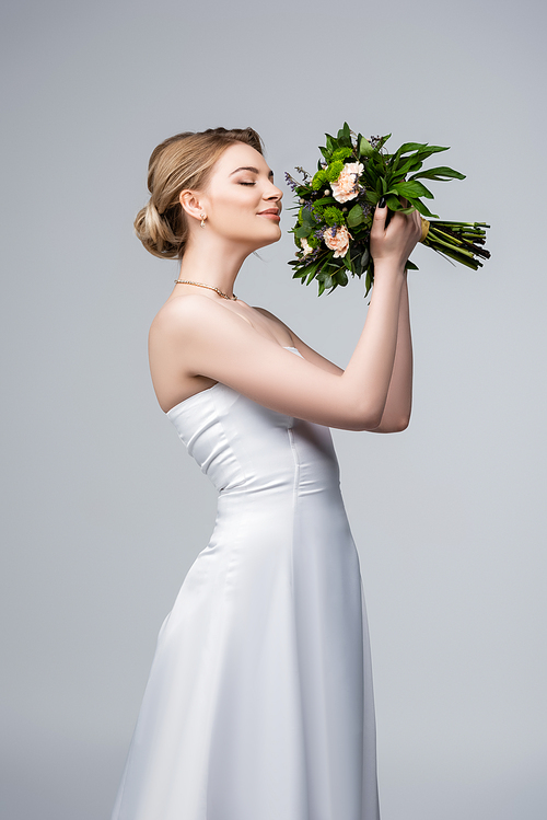 positive bride in white dress smelling wedding flowers isolated on grey