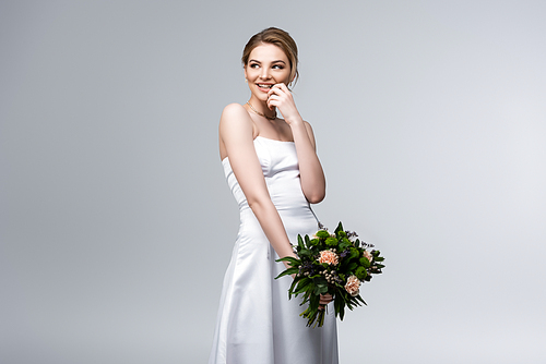 cheerful bride in white wedding dress holding flowers isolated on grey