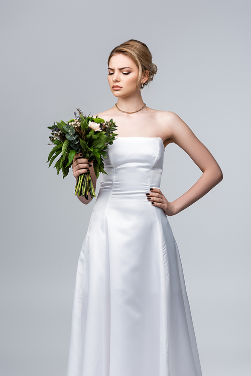 attractive bride in white dress looking at wedding flowers isolated on grey