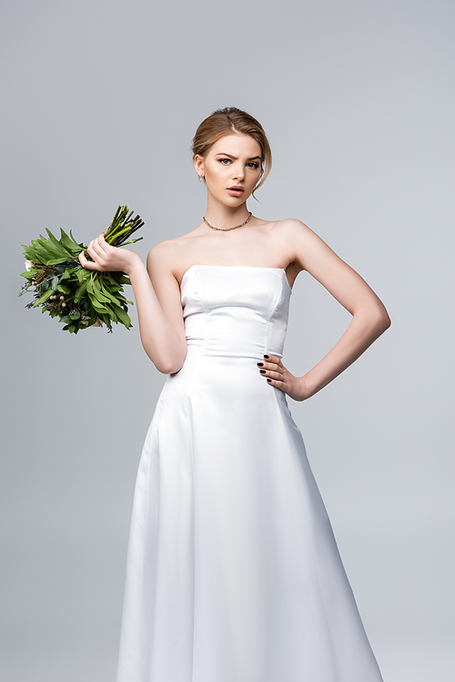 attractive bride in white dress holding wedding flowers and standing with hand on hip isolated on grey