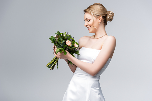 young and happy bride in white dress looking at bouquet of flowers isolated on grey