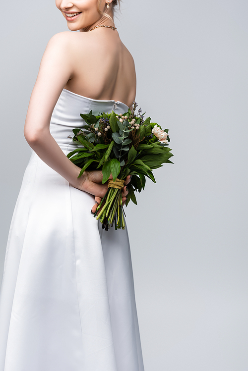 cropped view of young and cheerful woman in white wedding dress holding bouquet of flowers isolated on grey