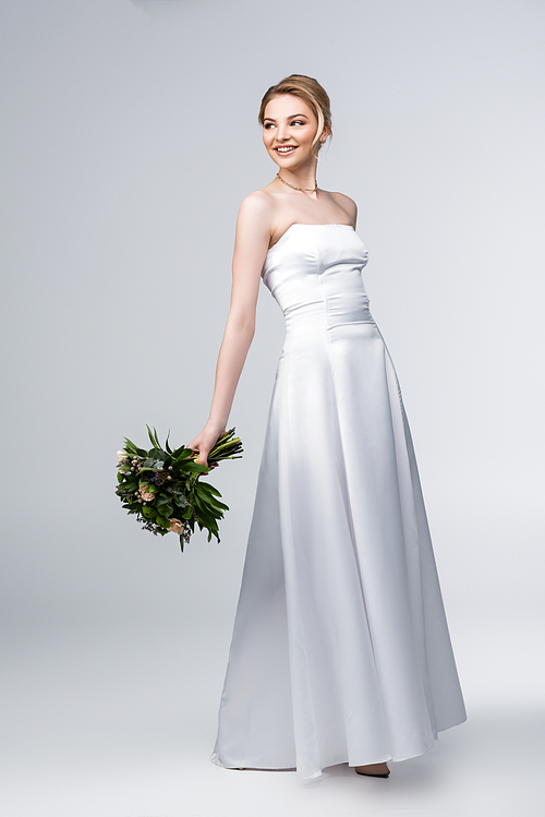 young and happy bride in white wedding dress holding bouquet of flowers on grey