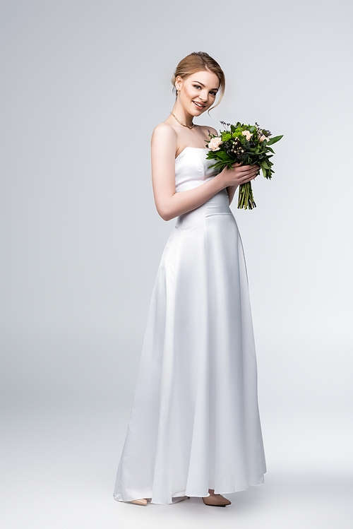happy bride in white wedding dress holding bouquet of flowers on grey
