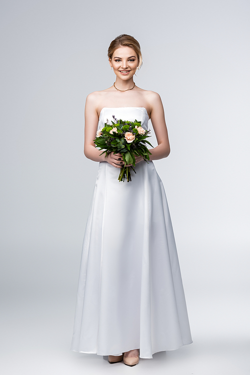smiling girl in white wedding dress holding bouquet of flowers on grey