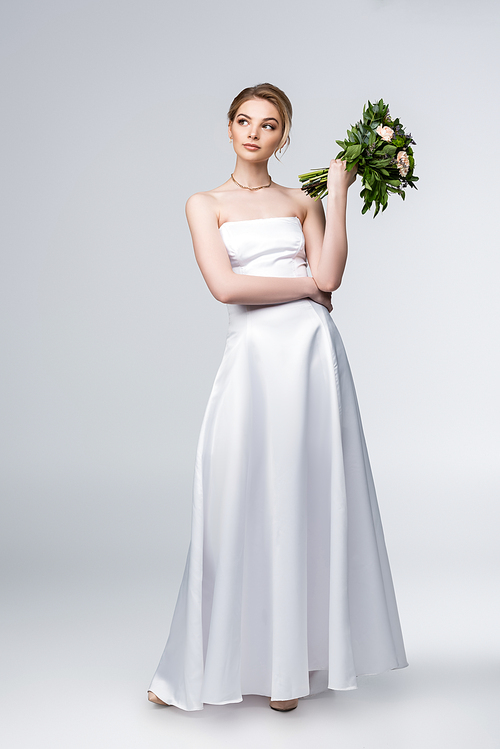 pensive girl in white wedding dress holding bouquet of flowers on grey