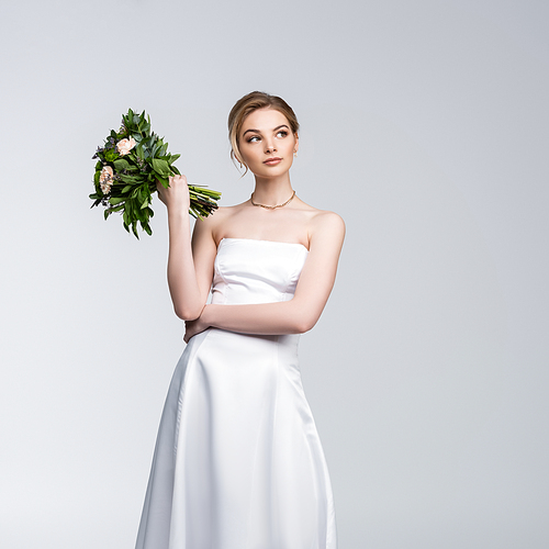pensive girl in white wedding dress holding bouquet of flowers isolated on grey
