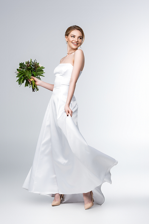 smiling bride touching white wedding dress and holding bouquet of flowers on grey