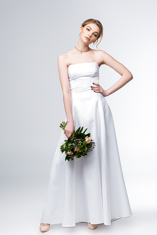 bride in elegant wedding dress holding bouquet of flowers and standing with hand on hip on grey