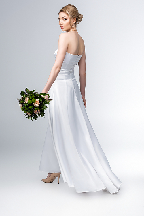 attractive bride in elegant wedding dress holding bouquet of flowers on grey