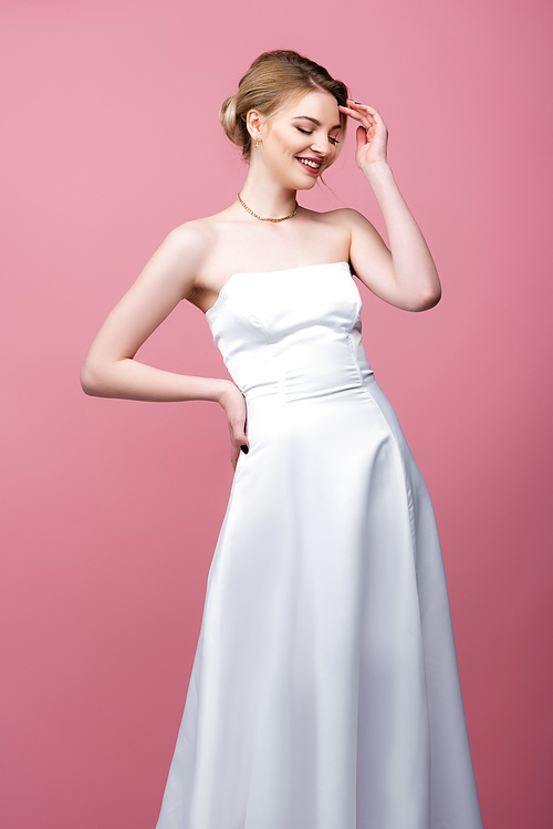 happy young bride in white wedding dress standing with hand on hip isolated on pink