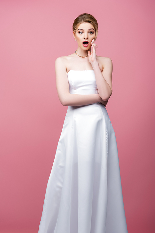 surprised bride in white wedding dress touching face isolated on pink