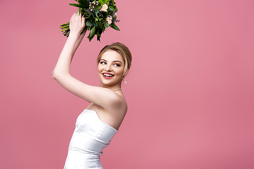 happy bride in white wedding dress holding flowers above head isolated on pink