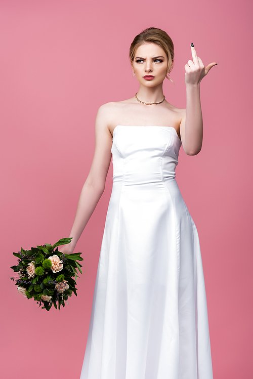 displeased bride in white wedding dress holding flowers and showing middle finger isolated on pink