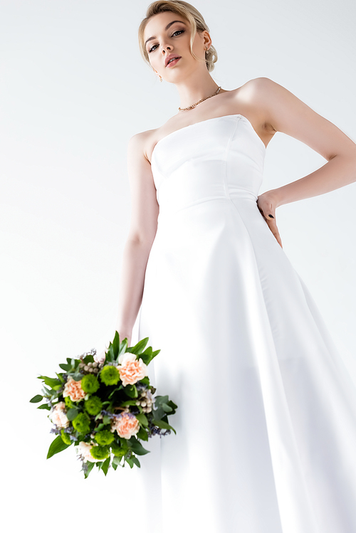 low angle view of elegant bride in wedding dress holding flowers and standing with hand on hip isolated on white