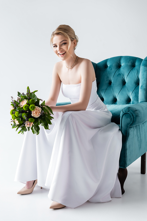cheerful bride in elegant wedding dress holding flowers while sitting in armchair on white