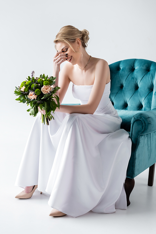 happy bride in elegant wedding dress holding flowers while sitting in armchair and touching face on white
