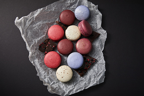 top view of assorted delicious colorful french macaroons on crumpled paper with chocolate on black background