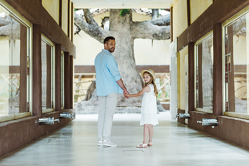 happy father holding hands with cheerful and cute daughter in straw hat and white dress while standing in zoo