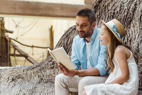 cheerful father reading book near cute daughter in straw hat near tree