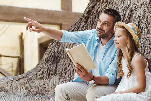 cheerful father holding book and pointing with finger near cute daughter in straw hat