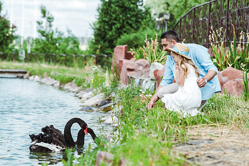 selective focus of cute kid in dress and straw hat sitting near happy father and lake with black swans