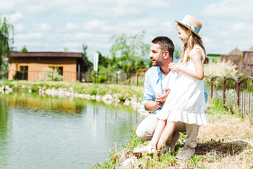 cute kid in dress and straw hat standing near happy father and lake