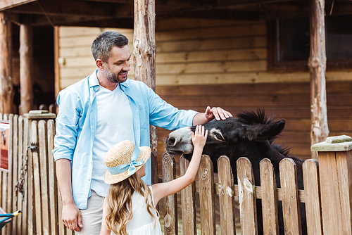 bearded man and cute daughter touching donkey while standing in zoo